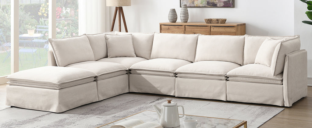 Spacious modular sectional sofa with ottoman in beige, arranged in a contemporary living room with wood accents and soft lighting.