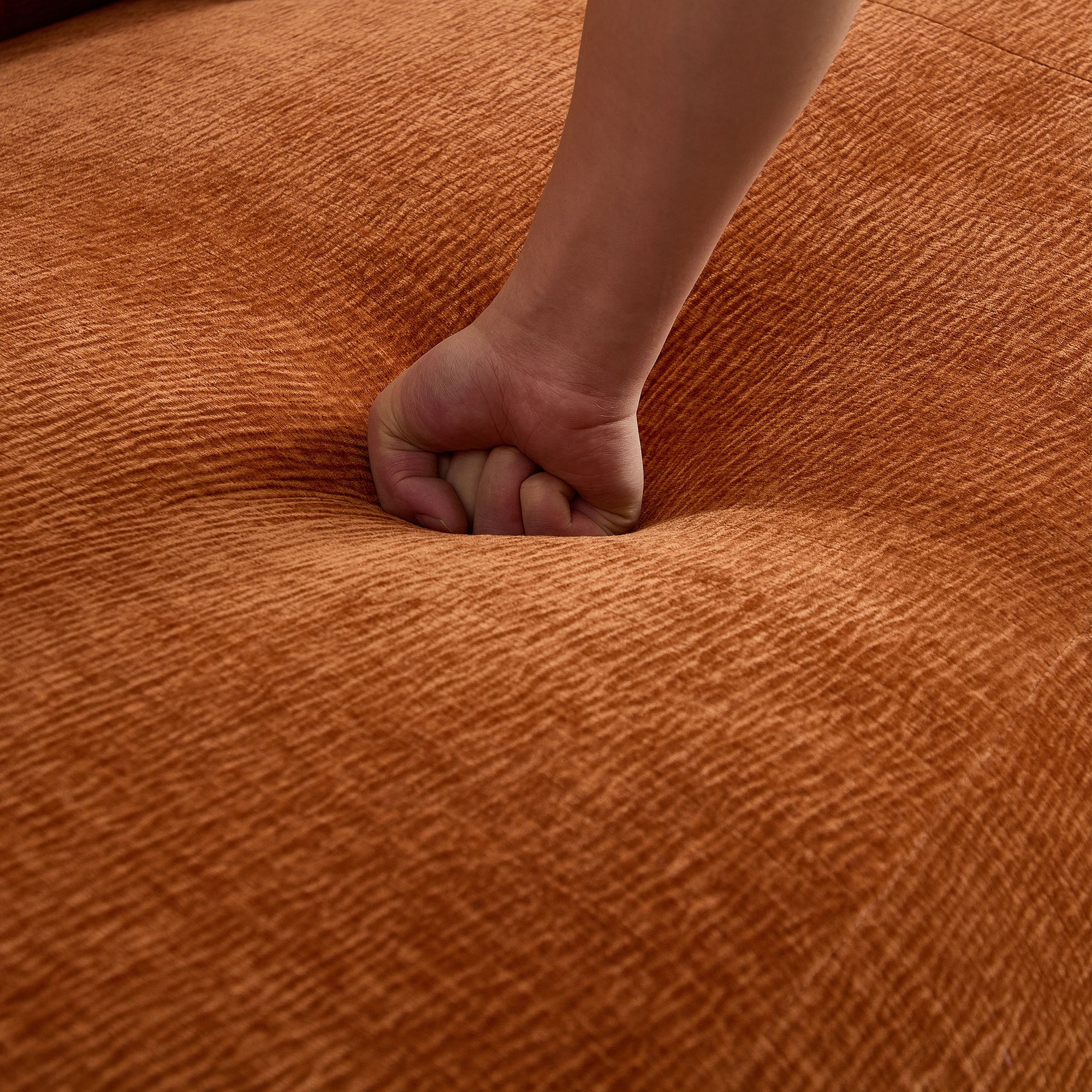 Hand pressing into the seat of the orange sectional sofa, showcasing the deep cushioning and soft fabric texture.