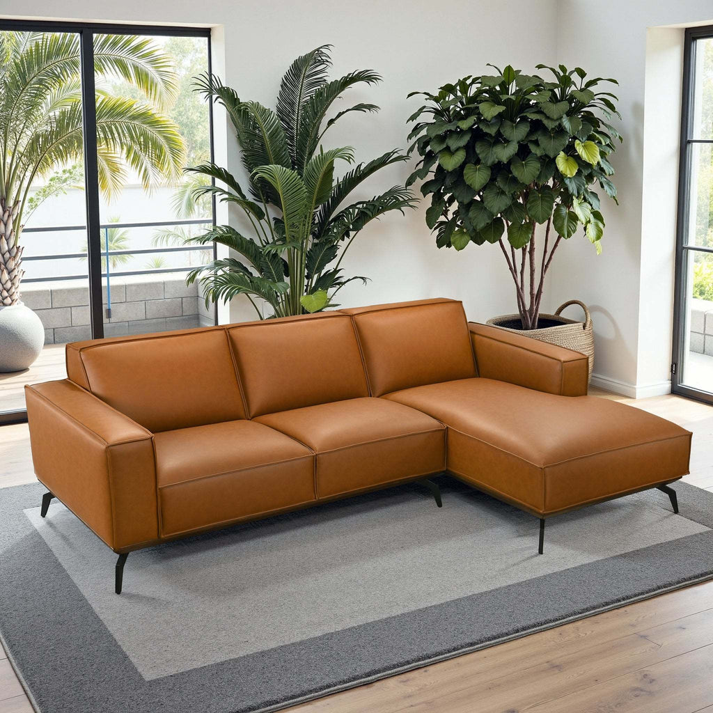 Modern tan leather right-facing sectional sofa with sleek black metal legs in a sunlit living room with lush greenery.