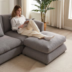 Woman lounging on gray upholstered ottoman of modular sofa, highlighting the comfort and relaxation features of the furniture.