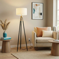 Modern tripod floor lamp with black metal legs, brass accents, and a cylindrical white fabric shade in a living room.