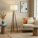 Modern tripod floor lamp with black metal legs, brass accents, and a cylindrical white fabric shade in a living room.