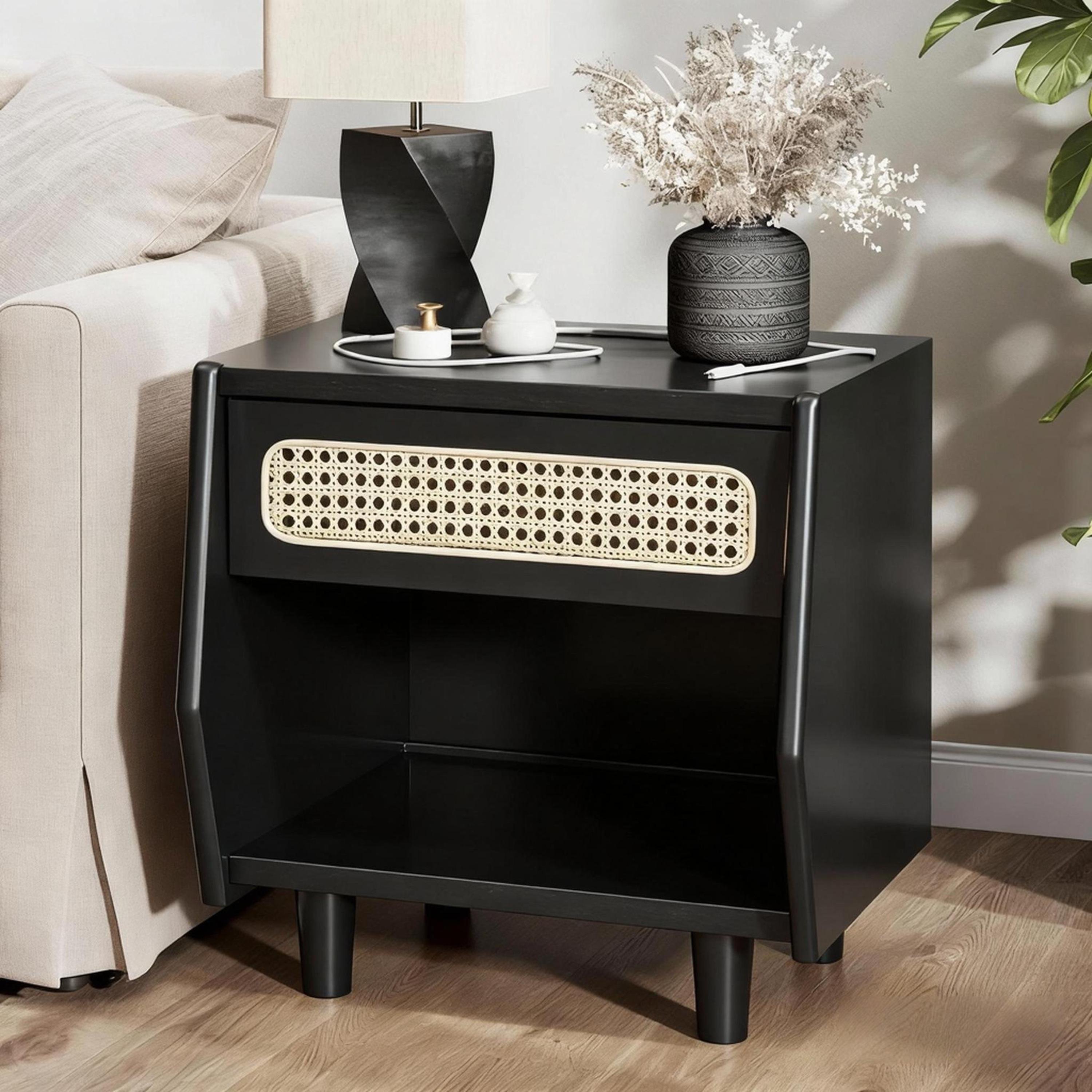 Modern black nightstand with rattan drawer detail, open shelf, and solid wood legs beside a beige sofa.