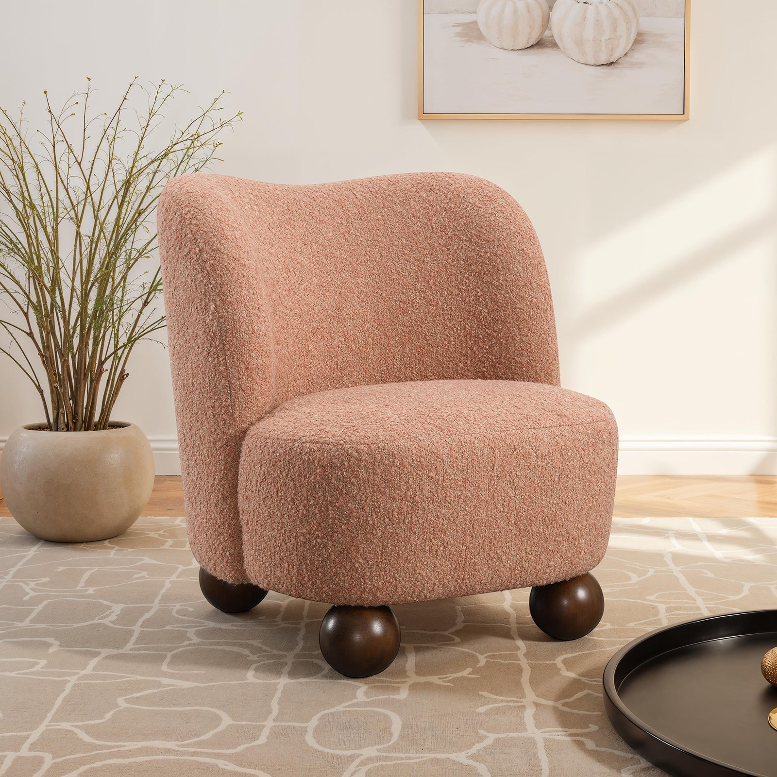 Pink accent chair with curved back in boucle fabric, styled in a modern living room with sculptural dark wood ball feet.