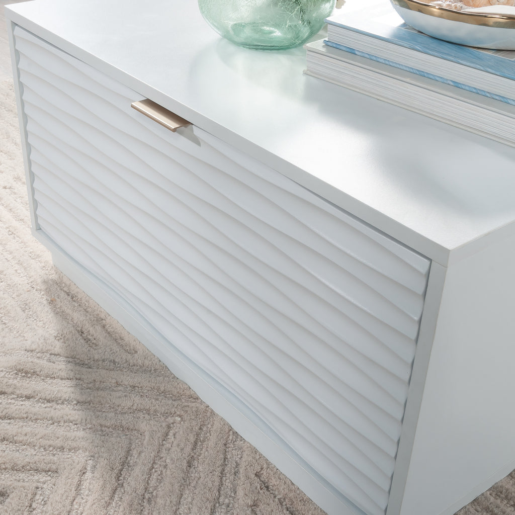 Angled view of the white coffee table with closed drawer, showcasing the ribbed texture and gold-tone handle in a coastal style.
