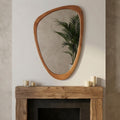 Irregular, pebble-shaped mirror with a smooth, yellowish-brown solid wood frame hanging above a rustic fireplace mantle.