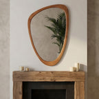 Irregular, pebble-shaped mirror with a smooth, yellowish-brown solid wood frame hanging above a rustic fireplace mantle.