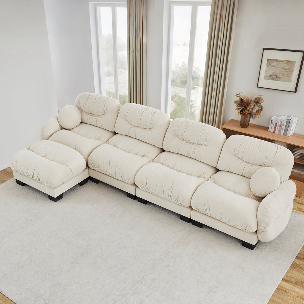 Beige 4-seater modular L-shaped sectional in pleated chenille fabric with curved arms, an ottoman, and two round pillows.