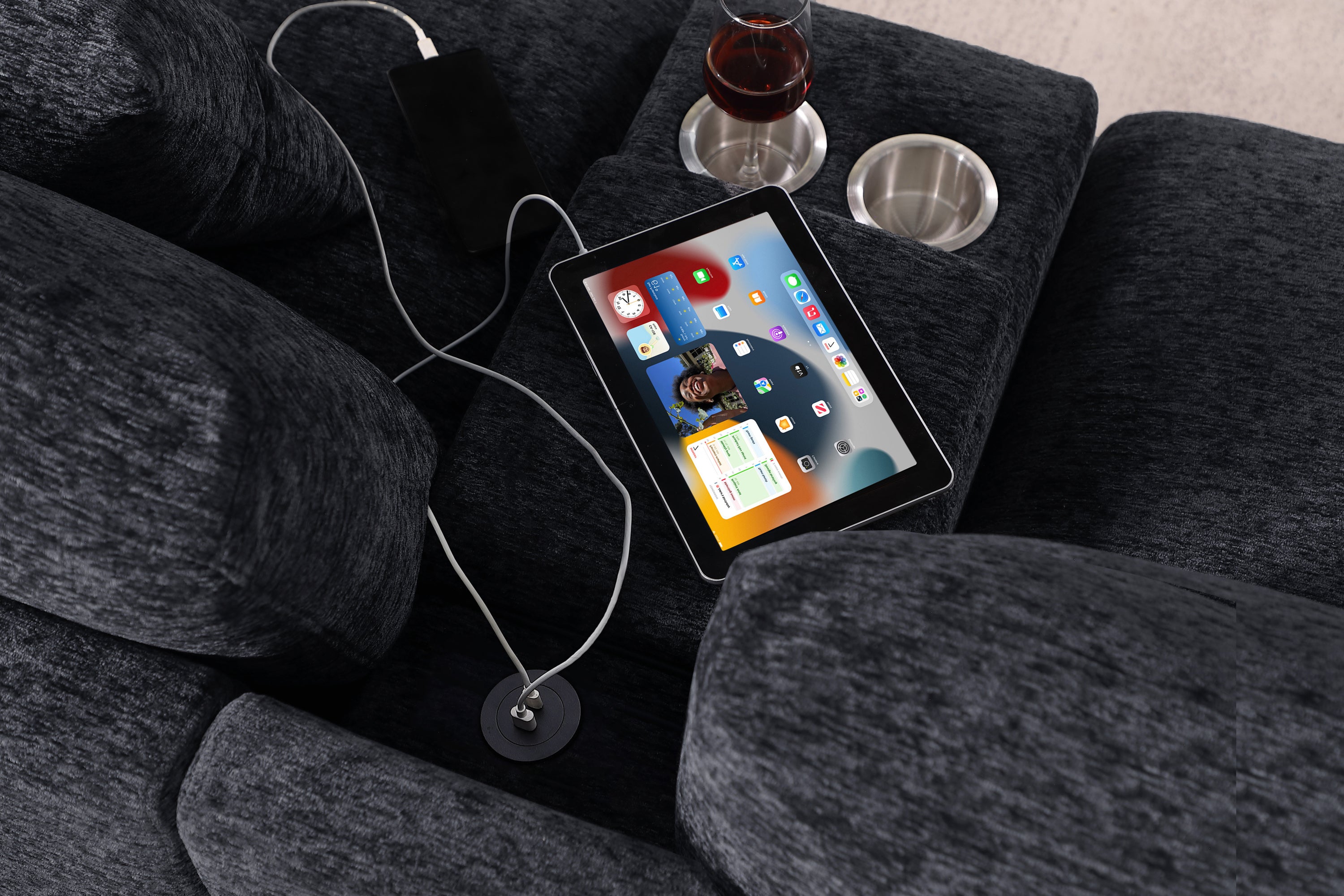 Close-up of Celine sectional sofa's USB charging port and cupholders with tablet and wine glass. Dark gray fabric and plush cushions.