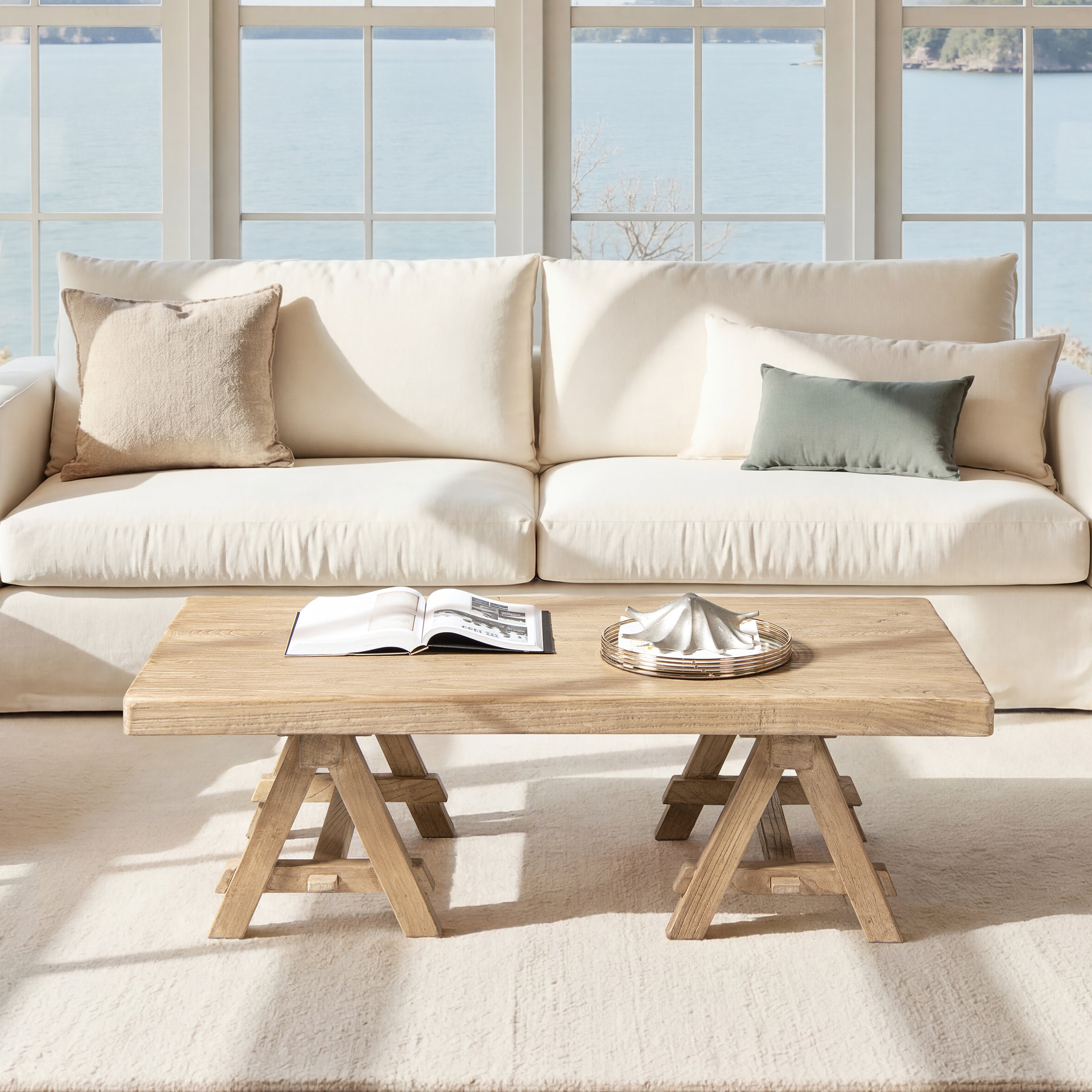 Rectangular coffee table in light natural brown reclaimed elm with a rustic trestle base, set before a white sofa.