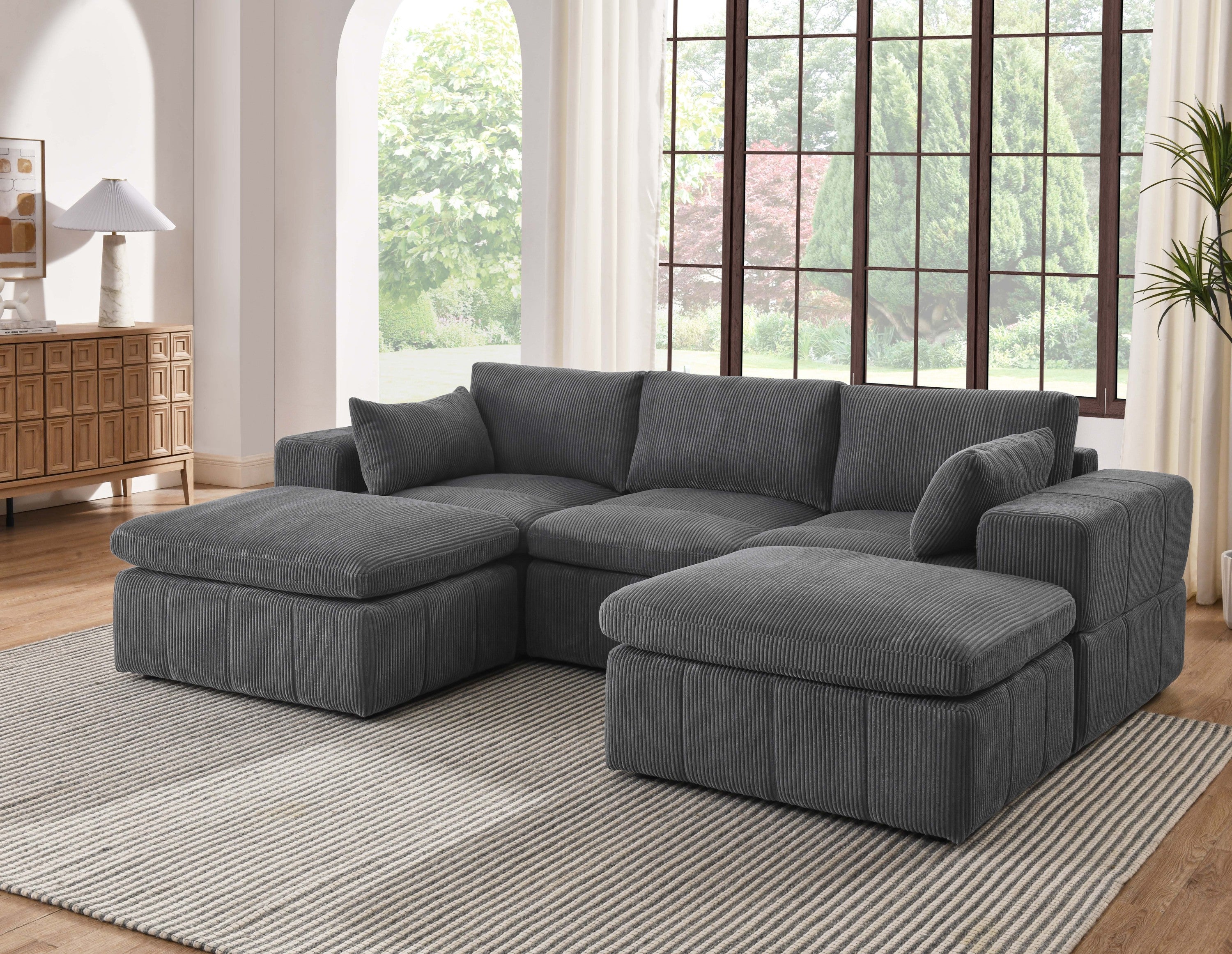 Modular sectional with ottomans in dark gray corduroy fabric placed in a bright living room. Features large arms and customizable layout.