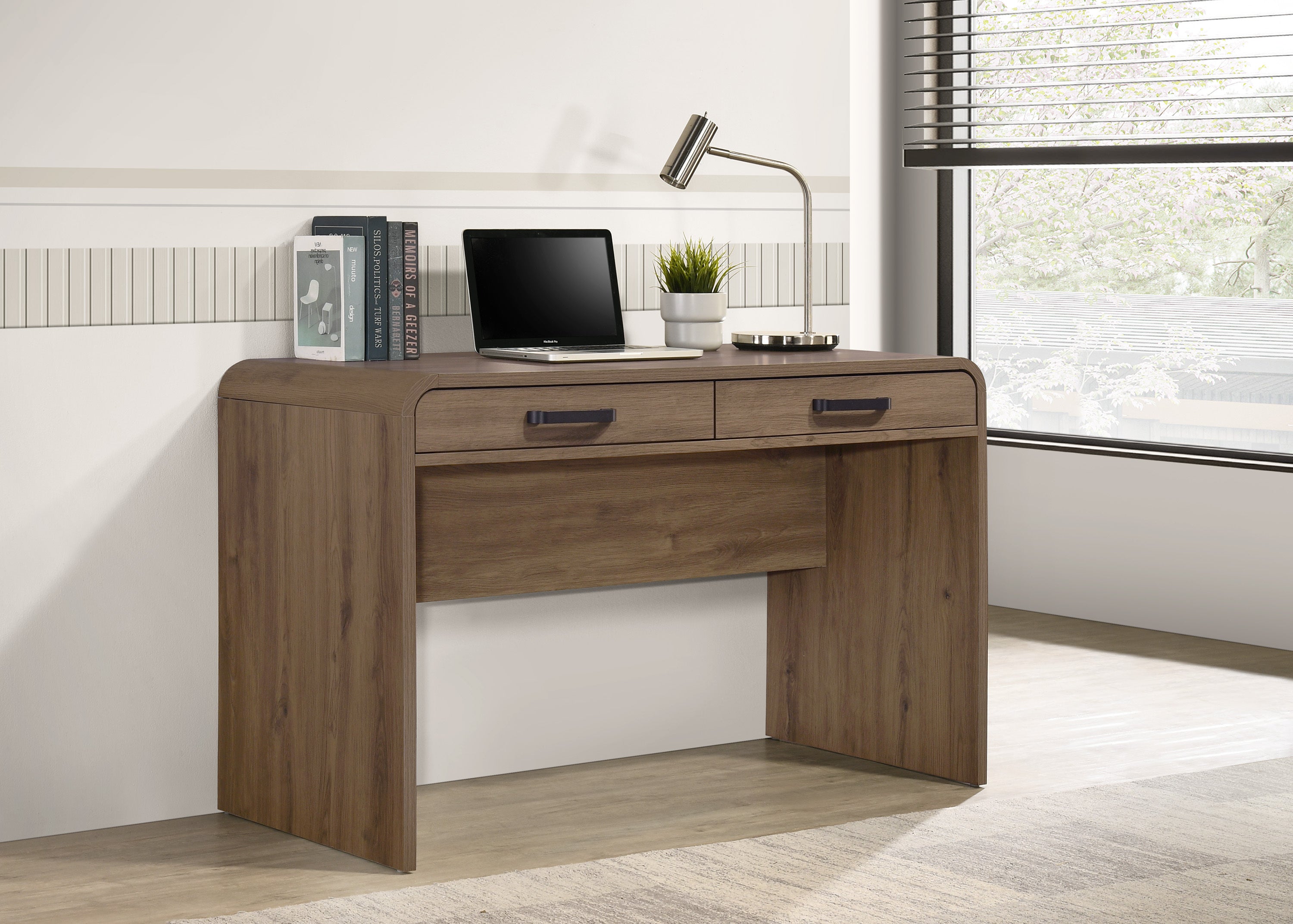 Modern light brown wooden writing desk with rounded corners, two drawers with black pulls, laptop, and desk lamp.