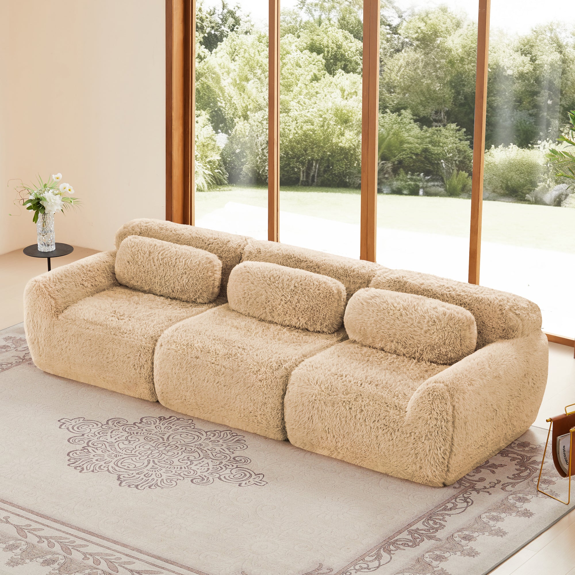 Modular sofa with plush fabric in beige, featuring three sections and back pillows, placed in a modern living room with large windows.