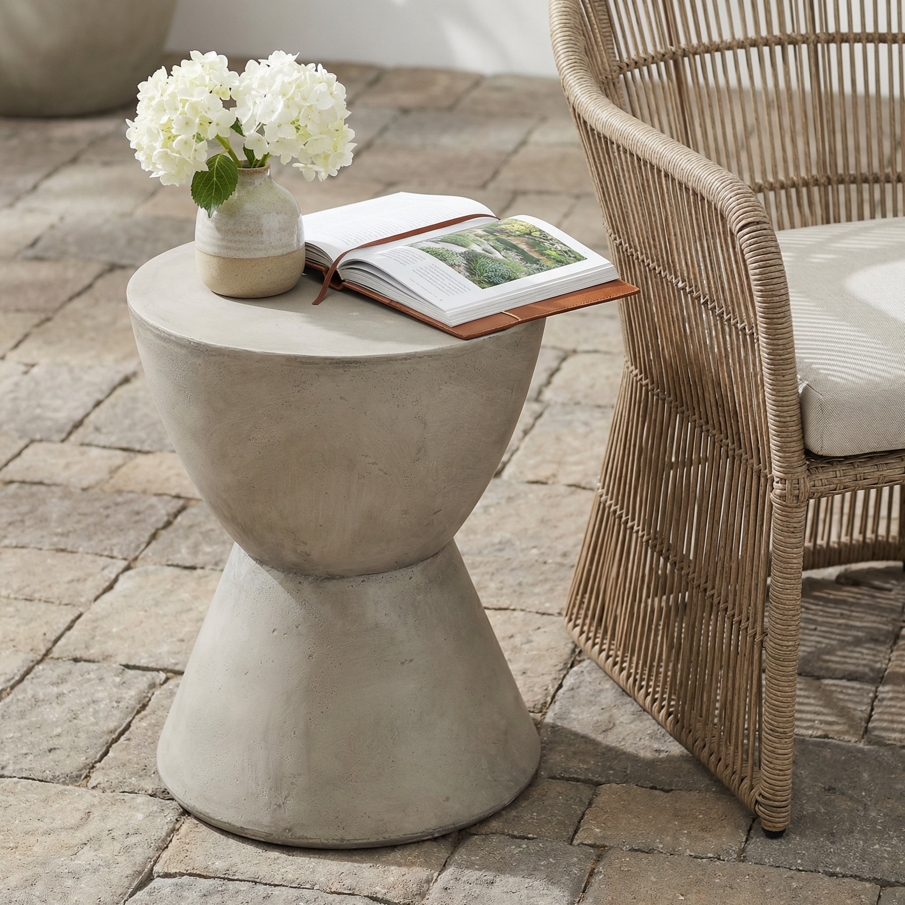 Light gray lightweight concrete outdoor side table with a modern, hourglass-shaped base on a stone patio.