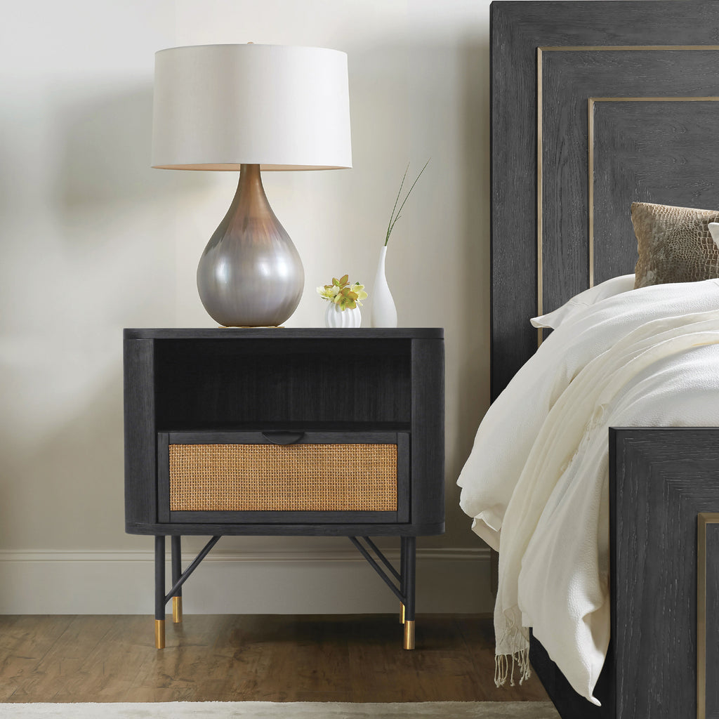 Black acacia nightstand with a rattan drawer, open shelf, and metal-tipped legs, styled in a modern bedroom.