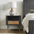 Black acacia nightstand with a rattan drawer, open shelf, and metal-tipped legs, styled in a modern bedroom.