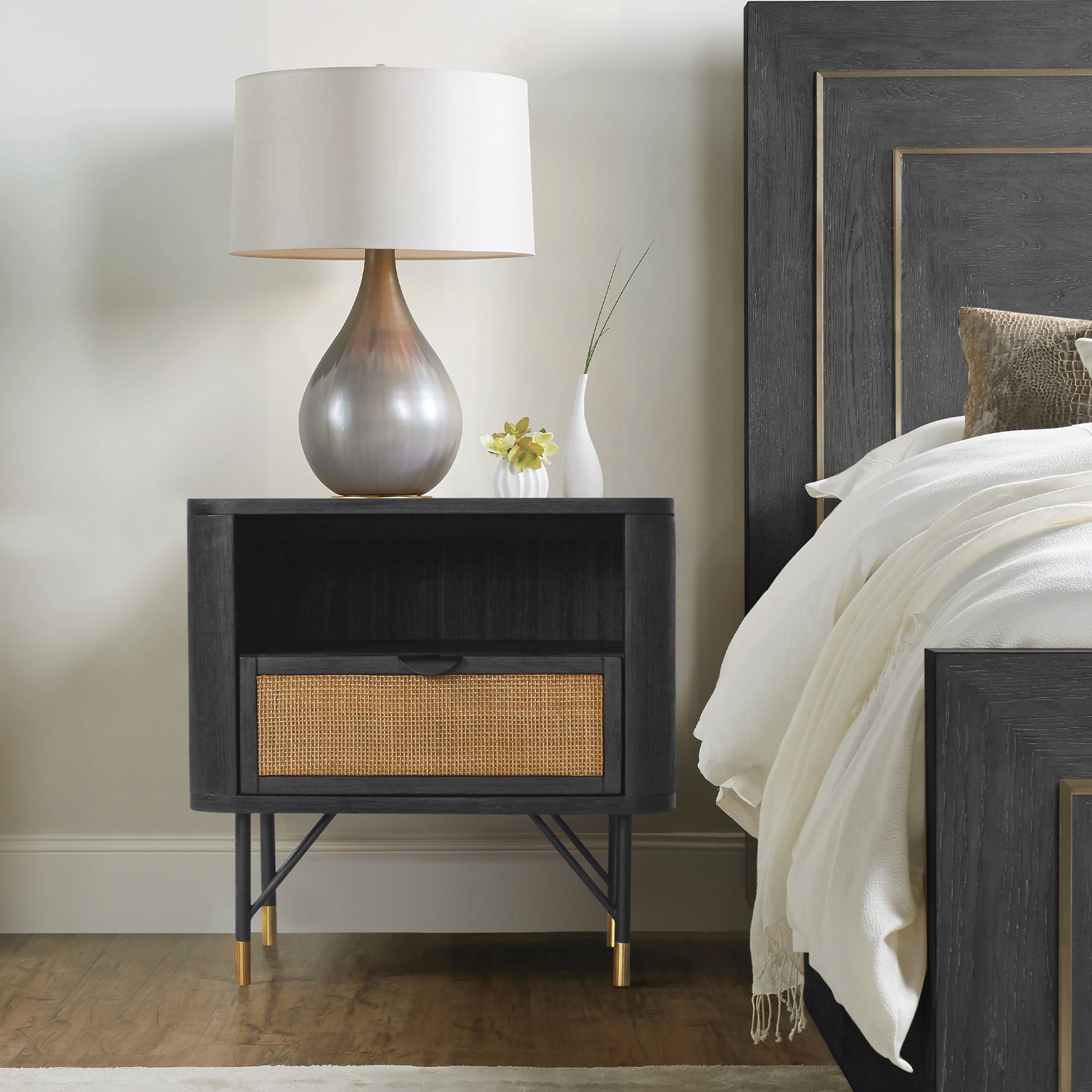Black acacia nightstand with a rattan drawer, open shelf, and metal-tipped legs, styled in a modern bedroom.