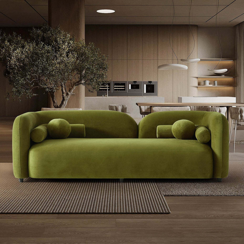 Olive green velvet Japandi sofa with a curved, low-profile silhouette and matching cylindrical bolster cushions.